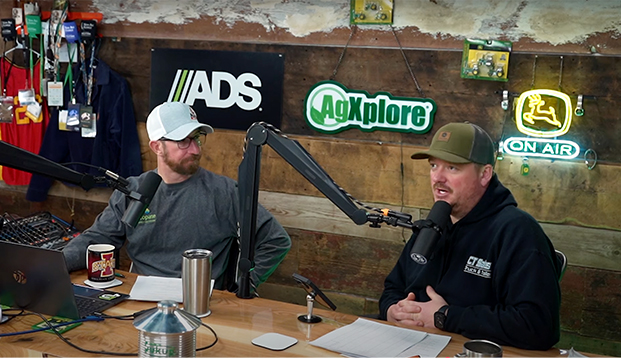 Two men talk on the set of a podcast. Signs for agriculture brands are on the wall behind them.